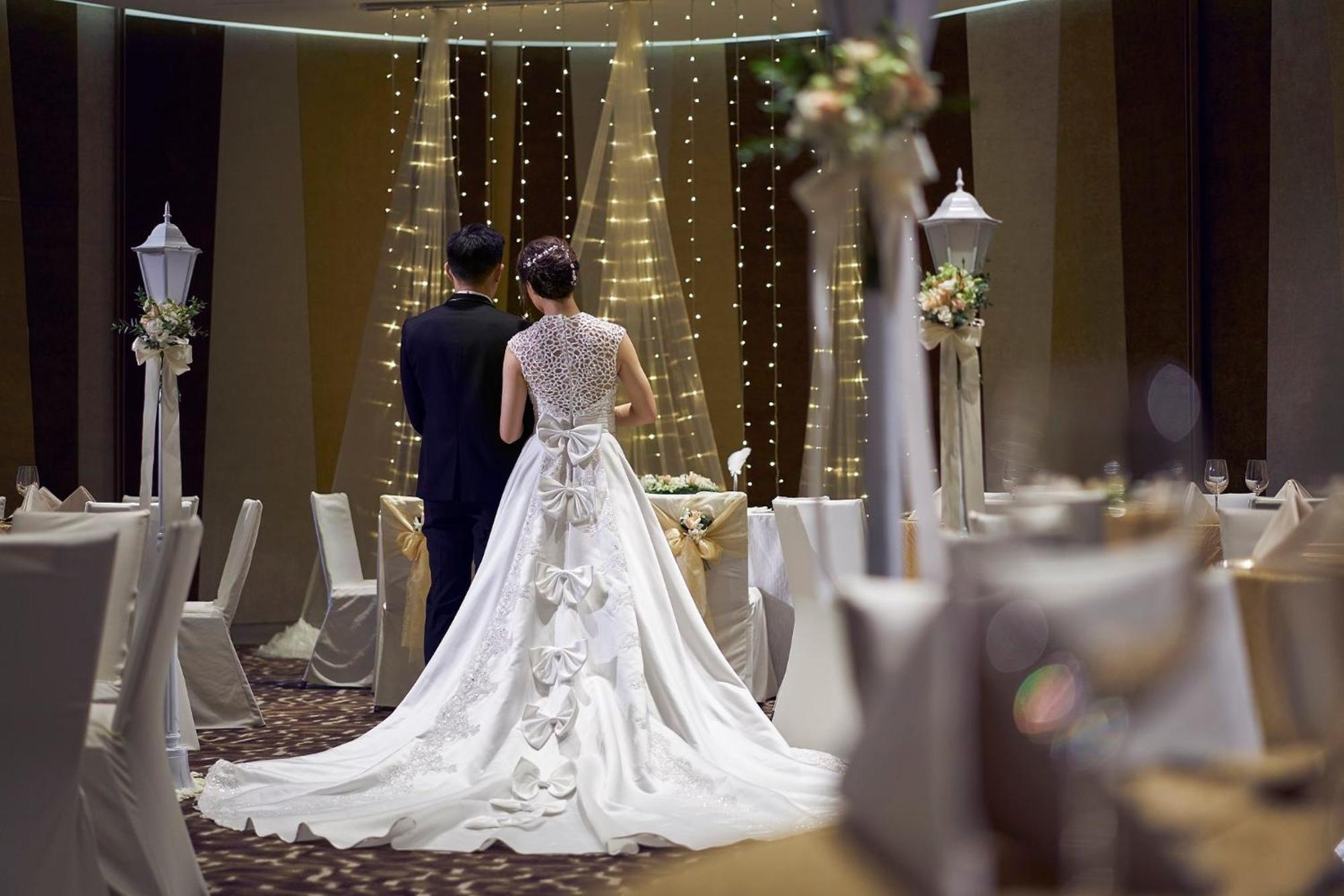 Crowne Plaza Changi Airport By Ihg Singapur Exterior foto The photo features a couple at a wedding venue. The bride, wearing an elegant white gown with intricate details and a long train, is seen from behind. The groom stands beside her, dressed in a dark suit. The background showcases a beautifully decorat